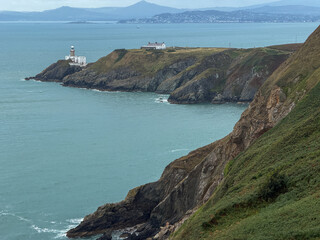 Baily Lighthouse stands on the rocky cliffs of Howth Head overlooking Dublin Bay, with the Irish coastline and distant mountains beneath a cloudy sky. © Ryan