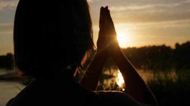 A woman holds her hands together in prayer against a luminous lakeside sunset. Backlight illuminates her silhouette, highlighting tranquil water reflections and serene evening air.