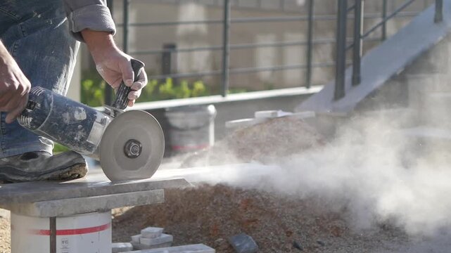 Professional worker cutting large stone tile using electric circular angle grinder with dust clouds
