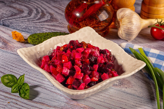 Diced beetroot salad with beans in white ceramic bowl on rustic wooden table surrounded by fresh garlic, tomato, basil, and pickles
