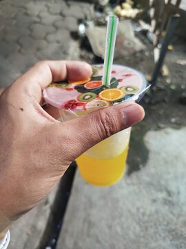 Refreshing colorful fruit drink in a plastic cup with a decorative lid and straw, held by hand outdoors, perfect for summer beverage and street food concepts.