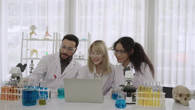 Group of happy men and women ethnic diversity, scientists and researchers wearing lab coats and goggles sitting and using a computer laptop for an online conference meeting together in the laboratory.