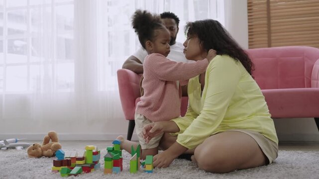 Family day concept. Happy African family is depicted with a mother and a cute little daughter playing with wooden blocks on the floor while the father sits on the sofa in the living room using tablet.