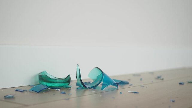 Broken blue and green glass pieces scattered on wooden floor near white wall