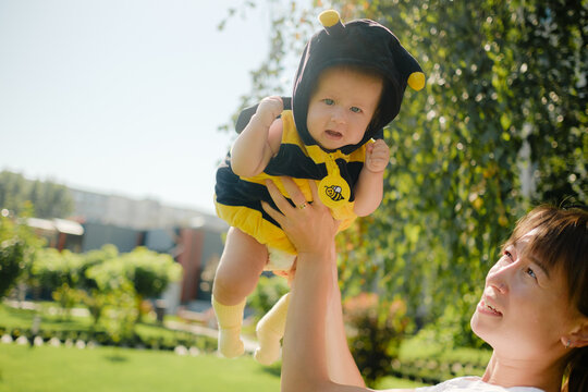 Mother holding baby in bee costume up in air outdoors in sunny park, happy mother, child care, freedom and childhood concept.