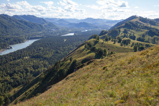 Panoramic river valley framed by rolling grassy hills and forested mountains, sunlit meadows and a winding river under a bright blue sky