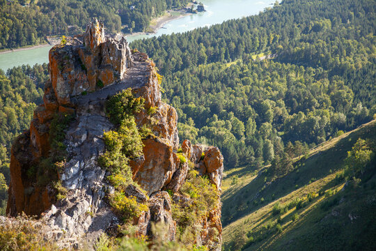 A rugged rocky cliff rises above a winding turquoise river and dense pine forest, bathed in warm sunlight. Dramatic panoramic viewpoint evokes adventure, Altai Devils Finger mount peak