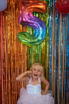 Joyful little girl celebrating fifth birthday with rainbow number five balloon
