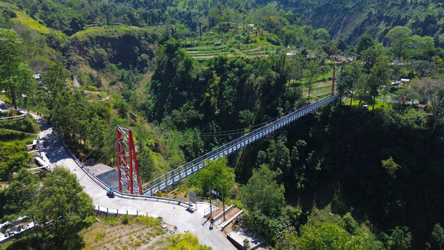 Suspension bridge girpasang, indonesia spans deep green valley. Red supports stand tall on either side. Road curves beneath the bridge gently