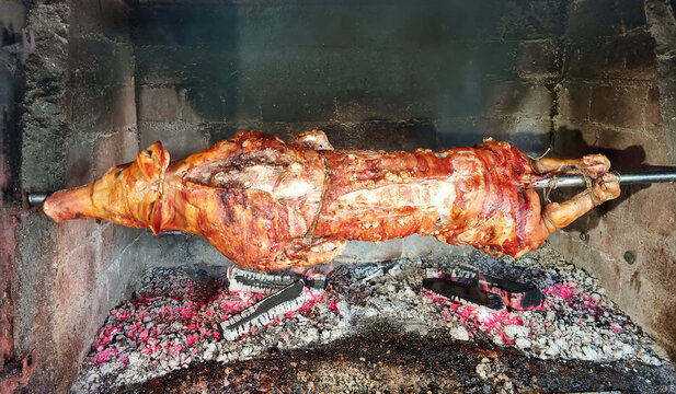 A whole pig turns slowly on a metal spit over glowing charcoal and wood embers inside a rustic stone fireplace.