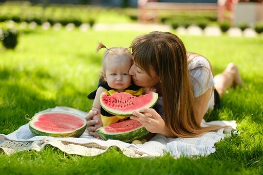 Happy mother and baby girl eating fresh red watermelon slice on green grass outdoors picnic, family summer concept.
