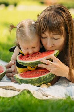 Happy mother and baby girl eating fresh red watermelon slice on green grass outdoors picnic, family summer concept.