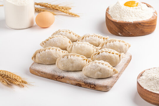 Raw uncooked Asian meat dumplings manti on wooden cutting board with flour and wheat ears on white table