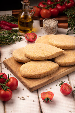 Raw uncooked breaded chicken meat cutlets on wooden board with herbs and cherry tomatoes on white wood table