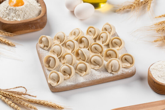 Raw uncooked pelmeni dumplings arranged on wooden board with flour eggs and wheat on light table