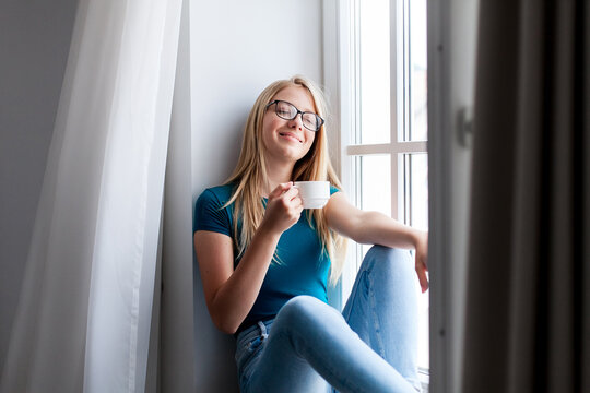 Young woman enjoying morning coffee, calming drink at home. Happy girl relaxing on windowsill. Concept of slow living, mindfulness, female mental health. Fresh air, wind from window
