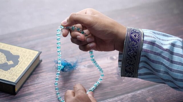 Man's hands counting prayer beads and holding Quran on wooden table