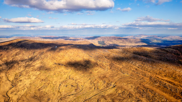 rolling hills covered in dry, golden grass under a blue sky with scattered clouds. Shadows from clouds cast across the textured, undulating landscape of hills and valleys.