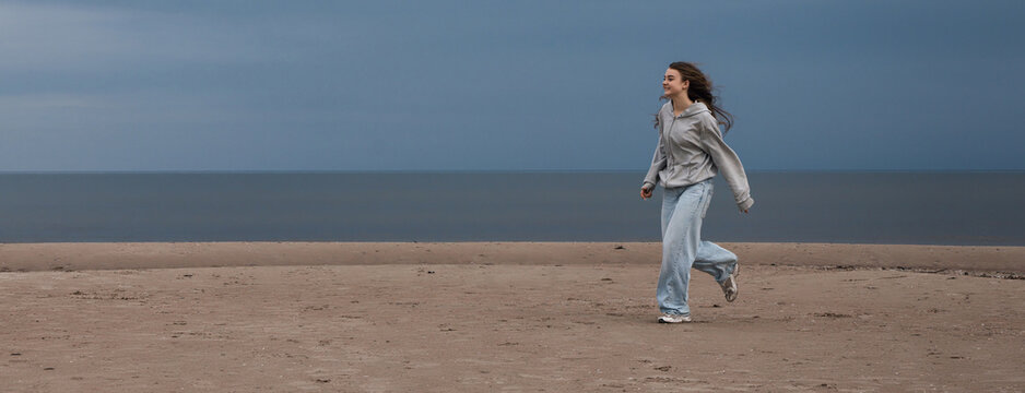 Smiling teenage girl walking on an empty beach on a windy overcast day with sea and copy space