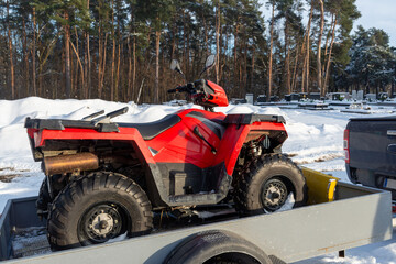 Red ATV quad bike stands on a metal utility trailer in a snow-covered outdoor area near a cemetery. Commercial image for off-road transport, vehicle hauling, trailer use, maintenance, winter equipment © Volha