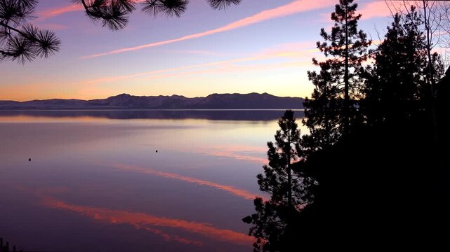 Stunning sunset over calm mountain lake with soft pink clouds and silhouette pine trees.