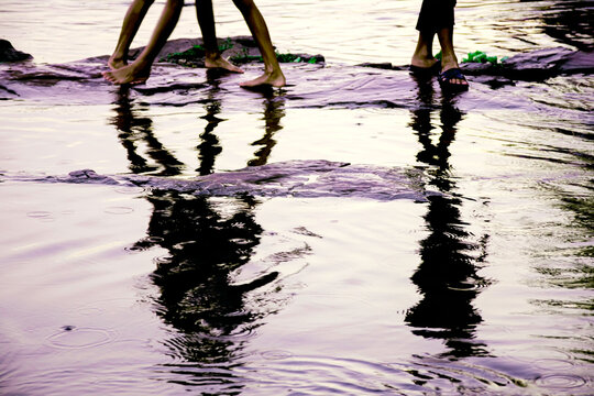 Low-angle view of people walking barefoot and in sandals across wet rocks near the water, with their silhouettes reflected in rippling puddles after rainfall