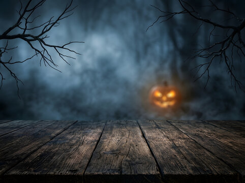 Spooky Halloween background with wooden table | Jack o lantern glowing in foggy forest at night | Rustic wood table surface for product display | Dark misty forest with creepy bare branches