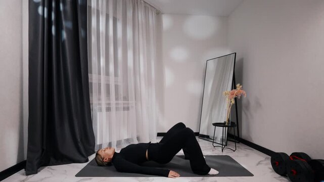 Young blonde woman in black activewear doing glute bridge during a home workout on a mat in a bright minimalist living room, side view