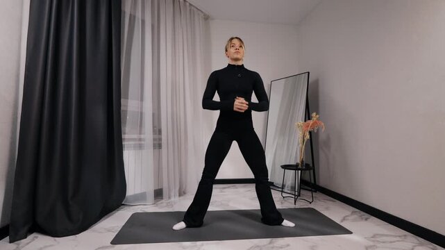 Young blonde woman in black activewear doing sumo squat during a home workout on a mat in a bright minimalist living room, front view