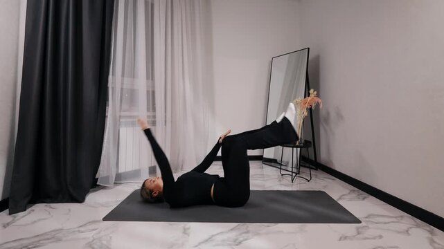 Young blonde woman in black activewear doing lying leg raise during a home workout on a mat in a bright minimalist living room, side view