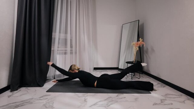 Young blonde woman in black activewear doing superman / back extension during a home workout on a mat in a bright minimalist living room, side view
