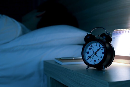 Vintage black alarm clock on a bedside table in a dark bedroom at night, with a person sleeping in the background, representing insomnia, early morning wakeup, or time management.