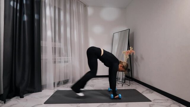 Young blonde woman in black activewear doing plank to downward dog with dumbbells during a home workout on a mat in a bright minimalist living room, side view