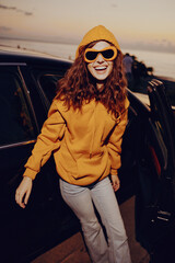 A smiling woman in an orange hoodie stands beside an open car door at sunset, conveying casual fashion, warmth, and carefree energy in a bright, outdoor moment. © SHOTPRIME STUDIO