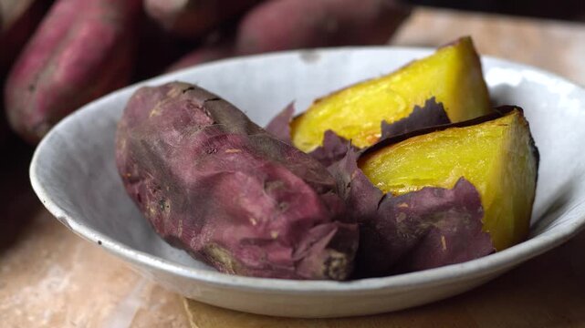 Delicious roasted Japanese sweet potatoes cut in half, showcasing their bright yellow flesh, served in a ceramic bowl with raw sweet potatoes in the background.