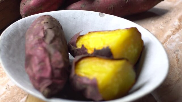 Delicious roasted Japanese sweet potatoes cut in half, showcasing their bright yellow flesh, served in a ceramic bowl with raw sweet potatoes in the background.