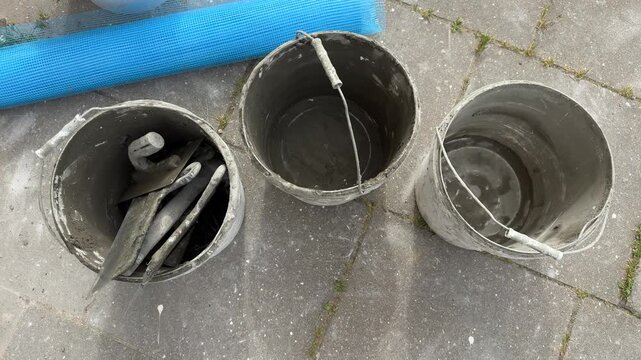 Three construction buckets with trowel tools and blue mesh roll on concrete pavement, outdoor work equipment, renovation, building materials, construction site