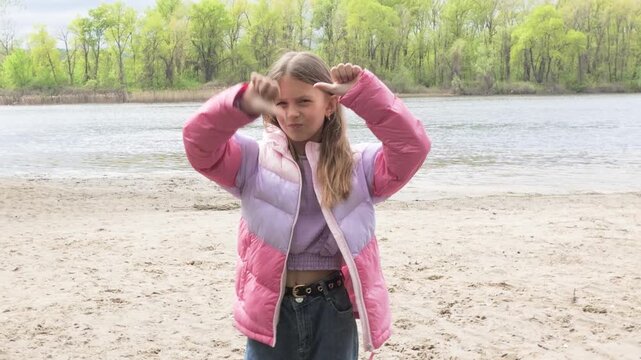 Young girl standing on a river beach wearing a pink jacket and showing a thumbs down gesture