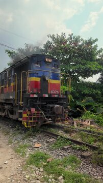 Vertical shot of a vintage locomotive restarting after a short stop, releasing thick smoke along the tracks. Lush green trees frame the colorful engine, contrasting with the industrial landscape.