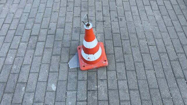 Damaged orange and white traffic cone on gray brick pavement, worn safety equipment with cracks and metal hook, outdoor urban street, construction zone marker, road work
