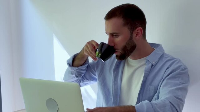 Young handsome man with a beard drinking morning coffee and working on a laptop at his desk, casually dressed while freelancing from home in a bright, minimalist apartment interior
