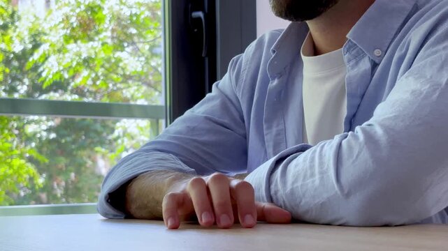 Worried young man sitting at a desk and tapping his fingers on the table, feeling anxious and stressed while looking out the window, showing impatience and concern about a problem