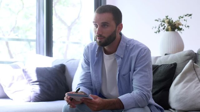 Thoughtful bearded man sitting on a couch in a bright living room, writing down ideas in a notebook before looking up and smiling confidently at the camera, feeling inspired and positive