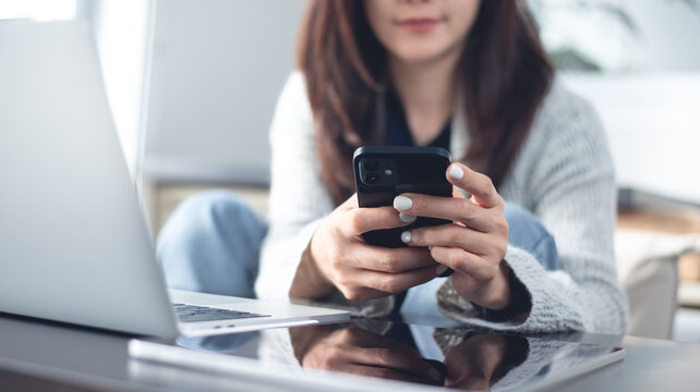 Asian woman sitting on the floor using smartphone during remote working at home with laptop on desk, digital work, online business concept