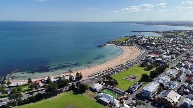 Aerial drone view of Williamstown Beach's curved sandy shoreline, blue calm bay waters, and coastal suburb with upscale residential houses. Melbourne, Victoria, Australia