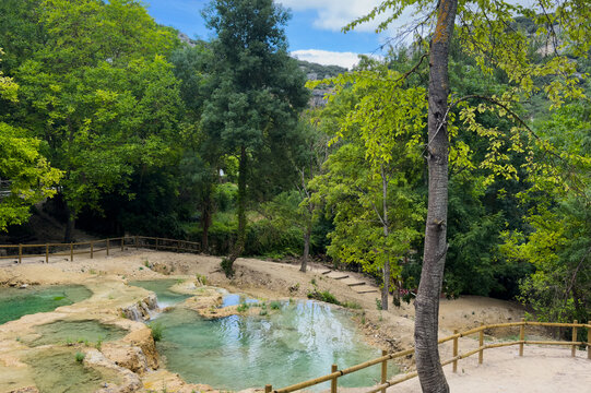 natural spring flowing over rocky terrain in a peaceful forest landscape, overlooking the serene riverside path with a wooden fence and green trees surrounding.