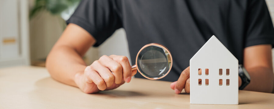 A man hand holds a magnifying glass inspecting a cottage house. Represents real estate appraisal, home inspection, and property assessment for search and investment