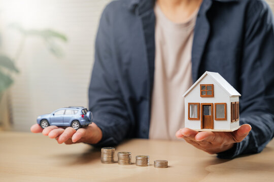 A person holds a car and house model above coin stacks, symbolizing real estate, auto loans, savings, and financial goals