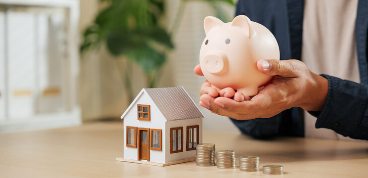 A close-up of hands with a piggy bank and house model, symbolizing saving and investment plans for property ownership. Highlights financial strategies, mortgage options