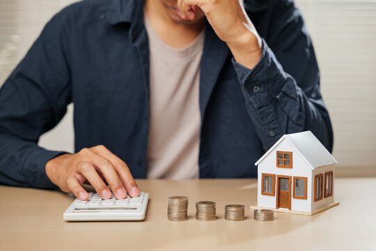 Stressed person holding a calculator, with stacks of coins and a house model nearby. Reflects financial planning challenges, tax concerns, and property calculations causing worry